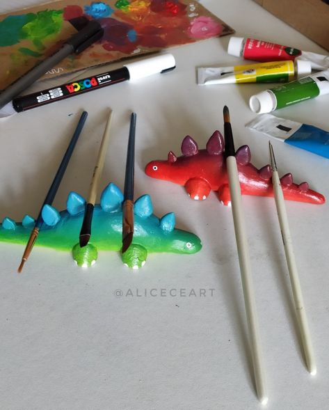 A photo of brightly coloured dinosaurs. They are made so that paintbrushes can be rested in the gaps between the spikes along the back. Around the dinosaurs are some paint tubes and a Posca paint pen Air Dry Clay Unique Ideas, Worm Paintbrush Holder, Clay Worm Paint Brush Holder, Clay Paint Holder, Paint Brush Rest Diy, Air Dry Clay Paint Pallet, Air Dry Clay Paintbrush Holder, Clay Paintbrush Rest, Polymer Clay Paint Brush Holder