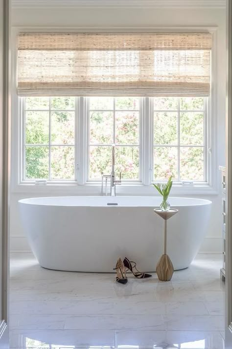 A bamboo roman shades hangs from a window located above an oval freestanding bathtub fitted with a polished nickel tub filler and placed on marble floor tiles behind a West Elm Faceted Brass Drink Table. Free Standing Bath In Front Of Window, Window Treatments Bathroom Master Bath, Classic Freestanding Bathtub, Big Window In Bathroom, Master Bath Freestanding Tub Ideas, Bathtub Windows, Tub In Front Of Window, Window Over Bathtub, Bathroom Window Treatments Over Tub