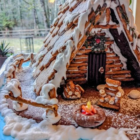 Gingerbread a-frame cabin on a lake with a candy campfire and mini chair and woodpile. To the side, a mini snowman and rosemary tree. The entrance to the pretzel log cabin is adorned with a thyme and candy wreath, and a jelly bean path. Pretzel Cabin Christmas, Ginger Bread Ideas Creative, Mountain Gingerbread House, Pretzel Gingerbread House Log Cabins, Gingerbread House With Rosemary, Log Cabin Pretzel House, Lake House Gingerbread House, Non Gingerbread House Ideas, Gingerbread Cabin Ideas