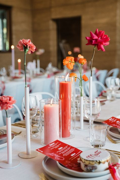 Vibrant wedding table setting with pink and red flowers, bold candles, and custom menus. A modern and colorful touch for a romantic reception. Pink And Orange Taper Candles, Coloured Candles Wedding Table, Colored Pillar Candles Wedding, Pink And Orange Table Setting, Indian Inspired Wedding, Red Table Settings, Coloured Pillar Candles, Wedding Reception Candles, Pillar Candles Wedding