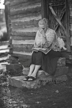 black and white photograph of a woman sitting on a log cabin porch reading a book