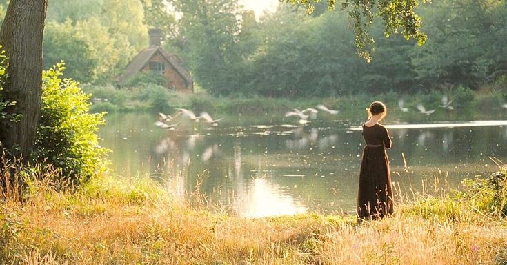 a woman standing in front of a body of water with birds flying above her head