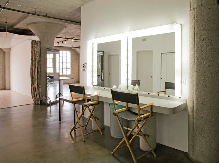 two chairs are sitting in front of a mirror with lights on it and mirrors hanging from the ceiling