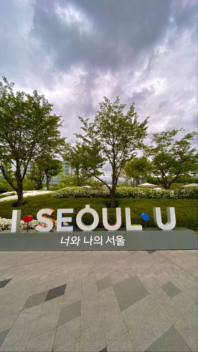 a sign that says seoul on it in front of some trees and bushes with clouds