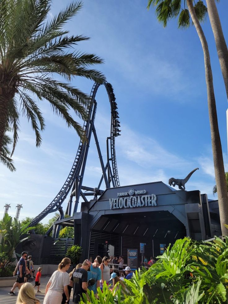 people are walking around in front of a roller coaster at the theme park, with palm trees and blue sky