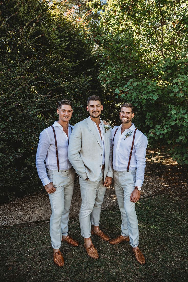 three men in white suits and brown suspenders standing next to each other with trees in the background