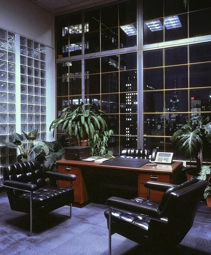 an office with two leather chairs and a desk in front of large windows at night