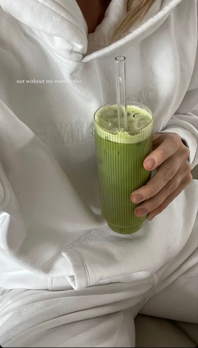 a woman in white hoodie holding a green smoothie and drinking it from a straw