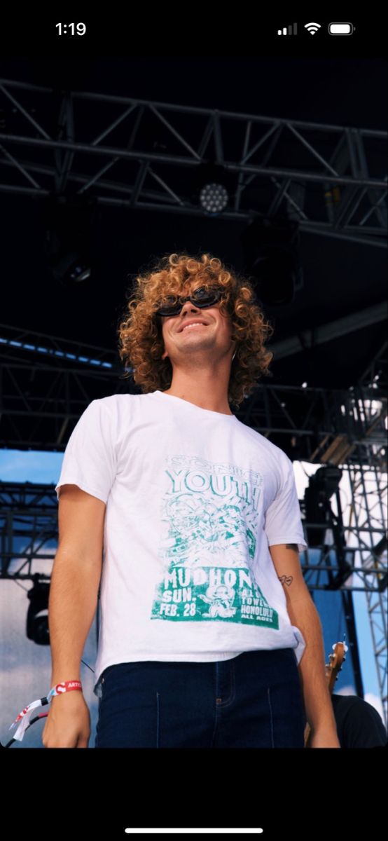 a man with curly hair and sunglasses standing in front of a stage wearing a white t - shirt