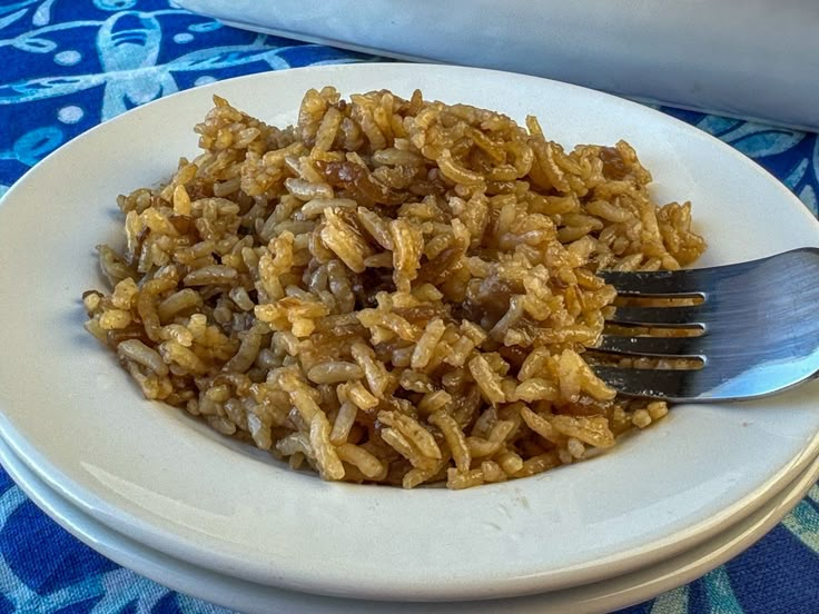 a white plate topped with rice next to a fork and bowl filled with food on top of a blue table cloth