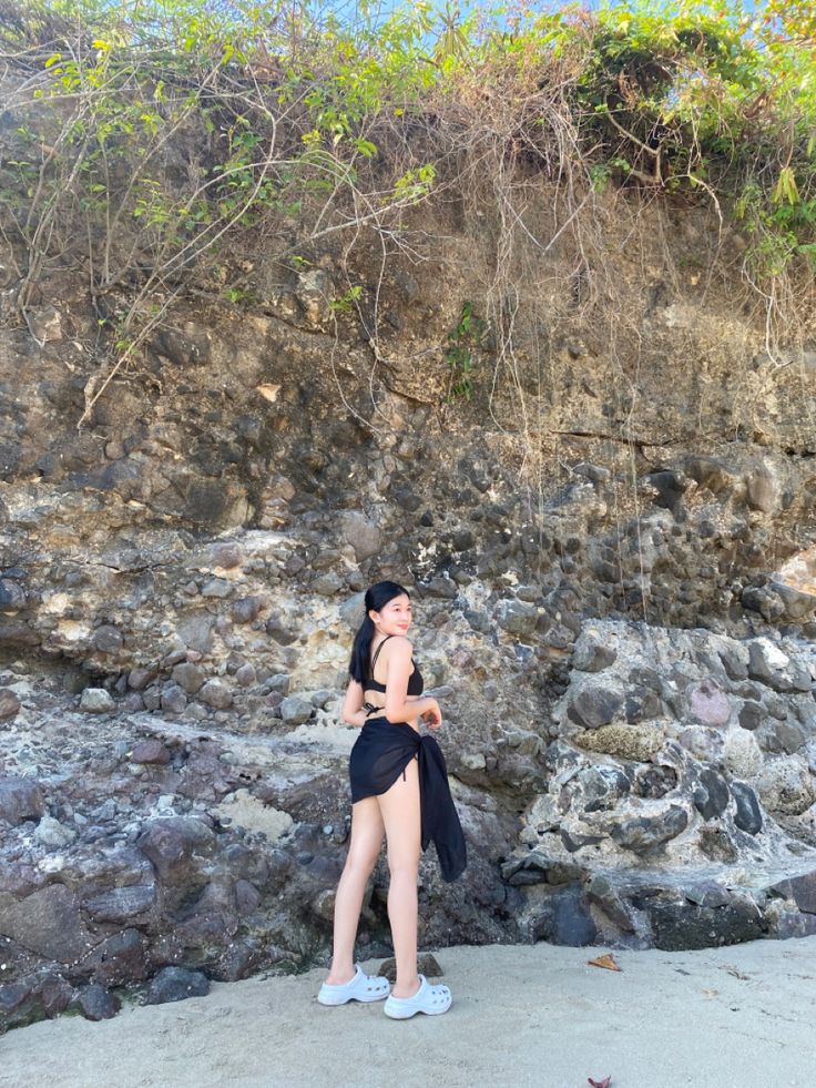 a woman in a black bathing suit standing next to a rock wall and looking up at the sky