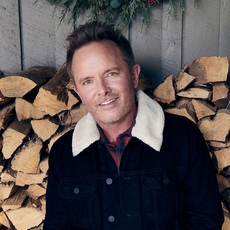 a man standing in front of a pile of firewood wearing a black jacket and white collared shirt