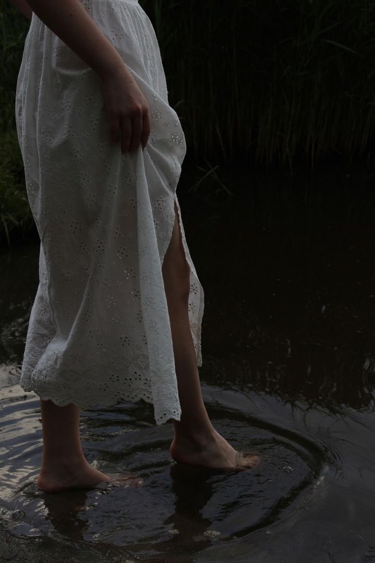 a woman is walking in the water wearing a white dress and holding her hand on her hip