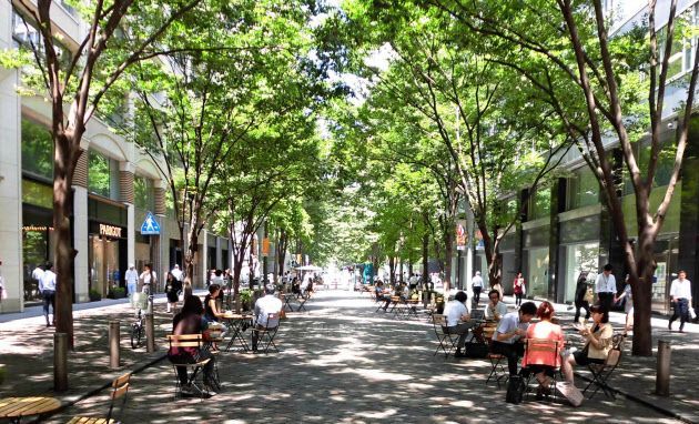 people are sitting on benches and walking down the sidewalk in an open area surrounded by trees