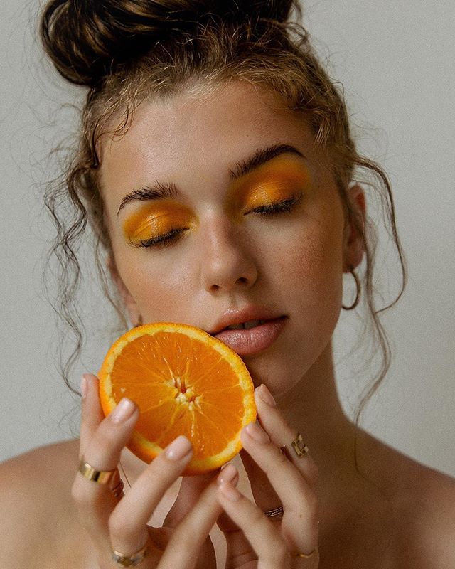 a woman holding an orange up to her face with yellow eyeshadow and gold makeup