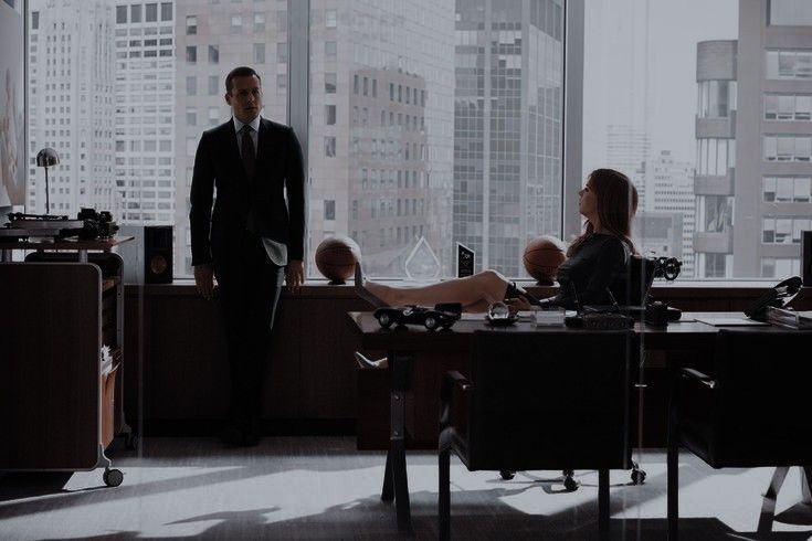 two people standing in an office looking out the window at another person sitting on a desk