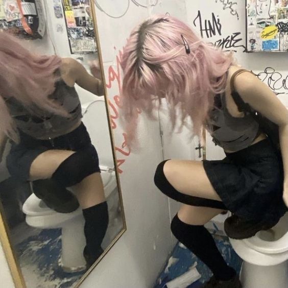 a woman with pink hair sitting on top of a toilet in a bathroom next to a mirror