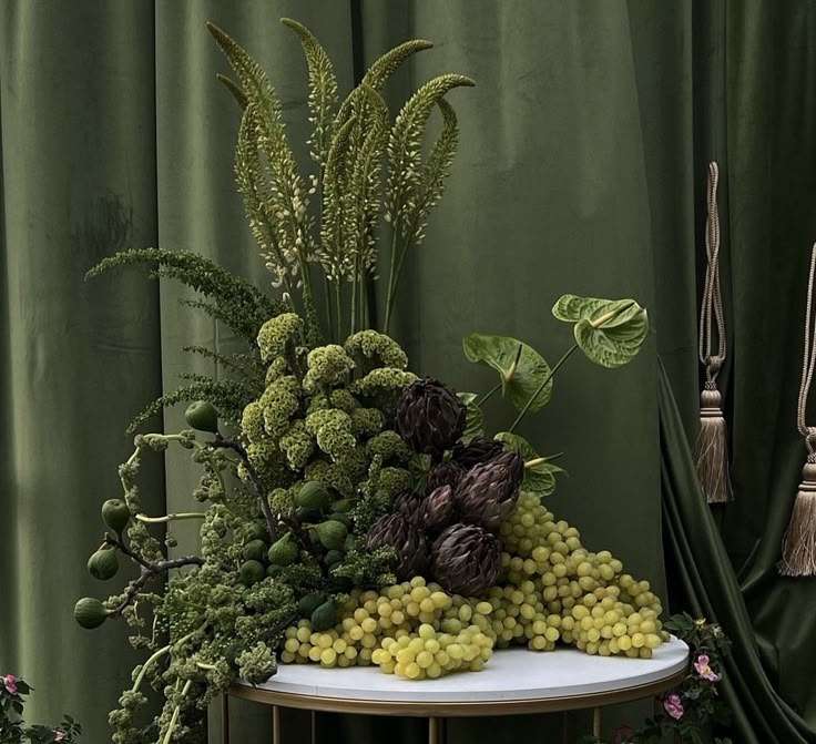 a table topped with lots of grapes and green plants next to a curtained window