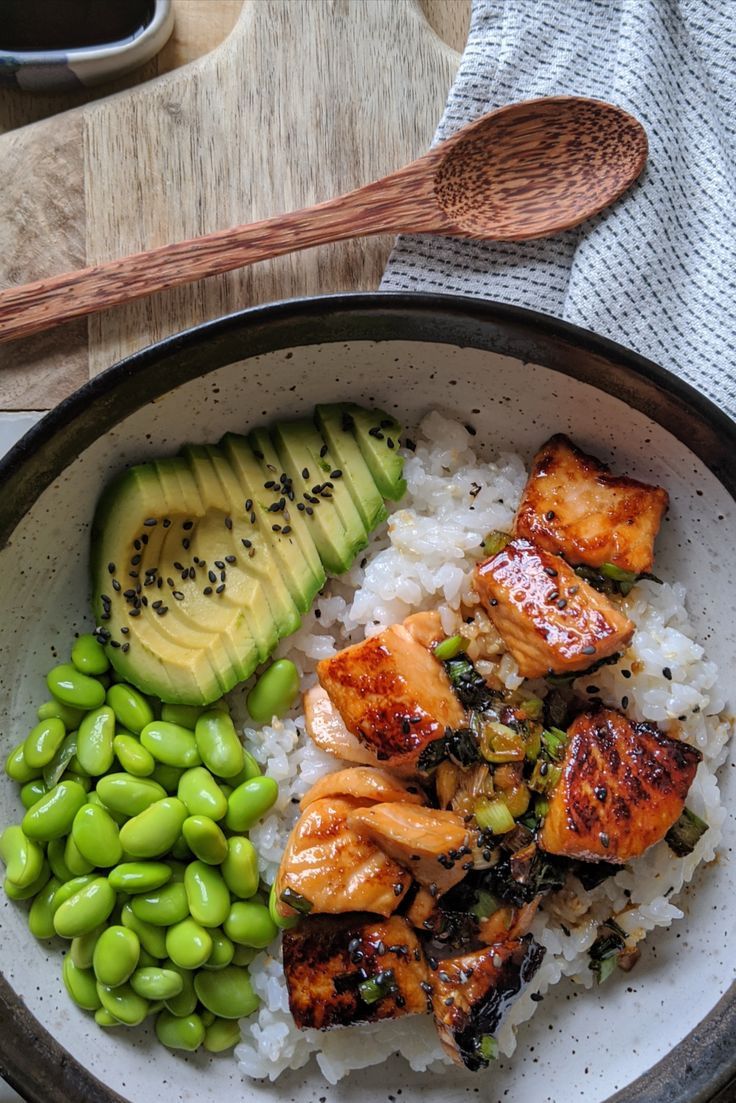 a bowl filled with rice, meat and vegetables next to a wooden spoon on top of a table