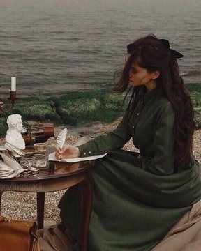 a woman sitting at a table writing on a piece of paper next to the ocean