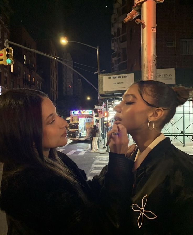 two women standing next to each other on a city street at night with traffic lights in the background