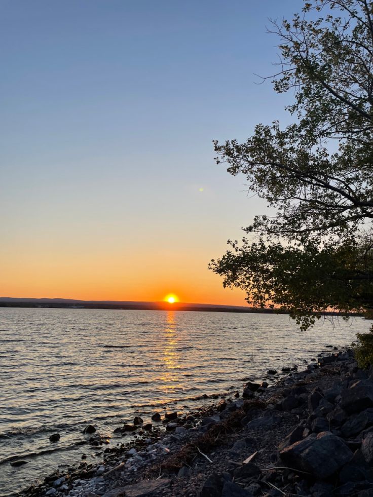 the sun is setting over the water and rocks on the shore with trees in the foreground