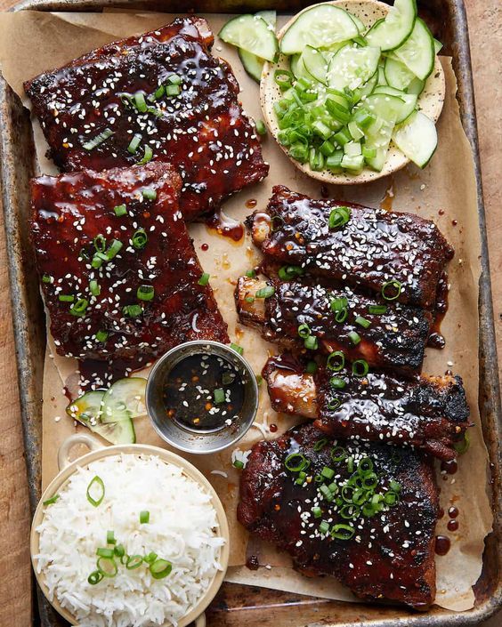 ribs, rice and cucumbers on a tray with dipping sauce in the middle