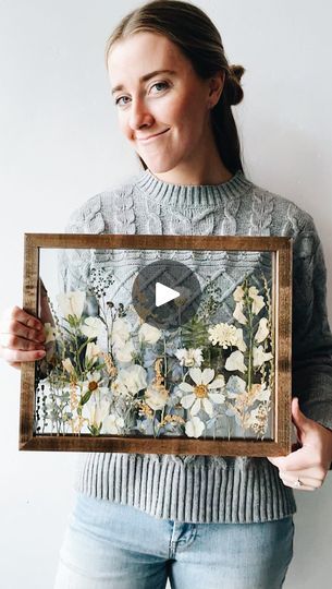 a woman holding up a framed painting with flowers on it and an arrow in the middle