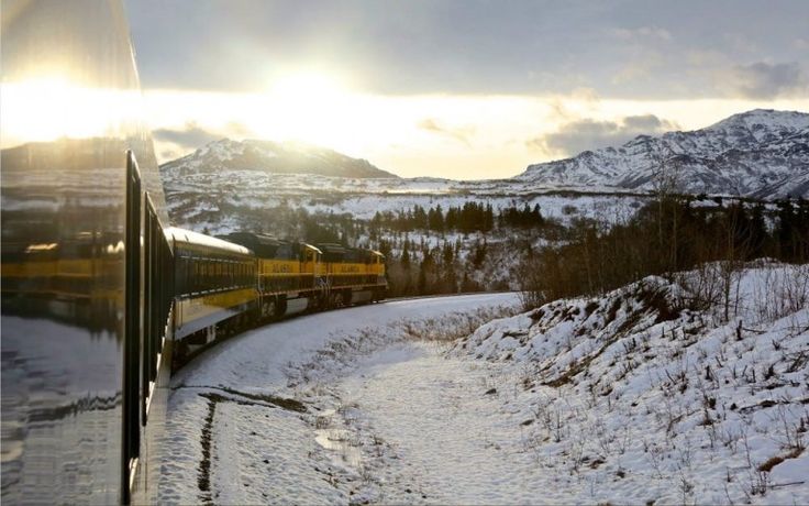 a train traveling down tracks next to snow covered hills and mountains in the distance with sun shining through clouds