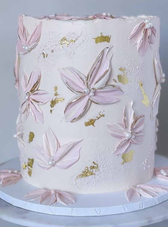 a white cake with pink and gold flowers on it sitting on a stand next to a wall