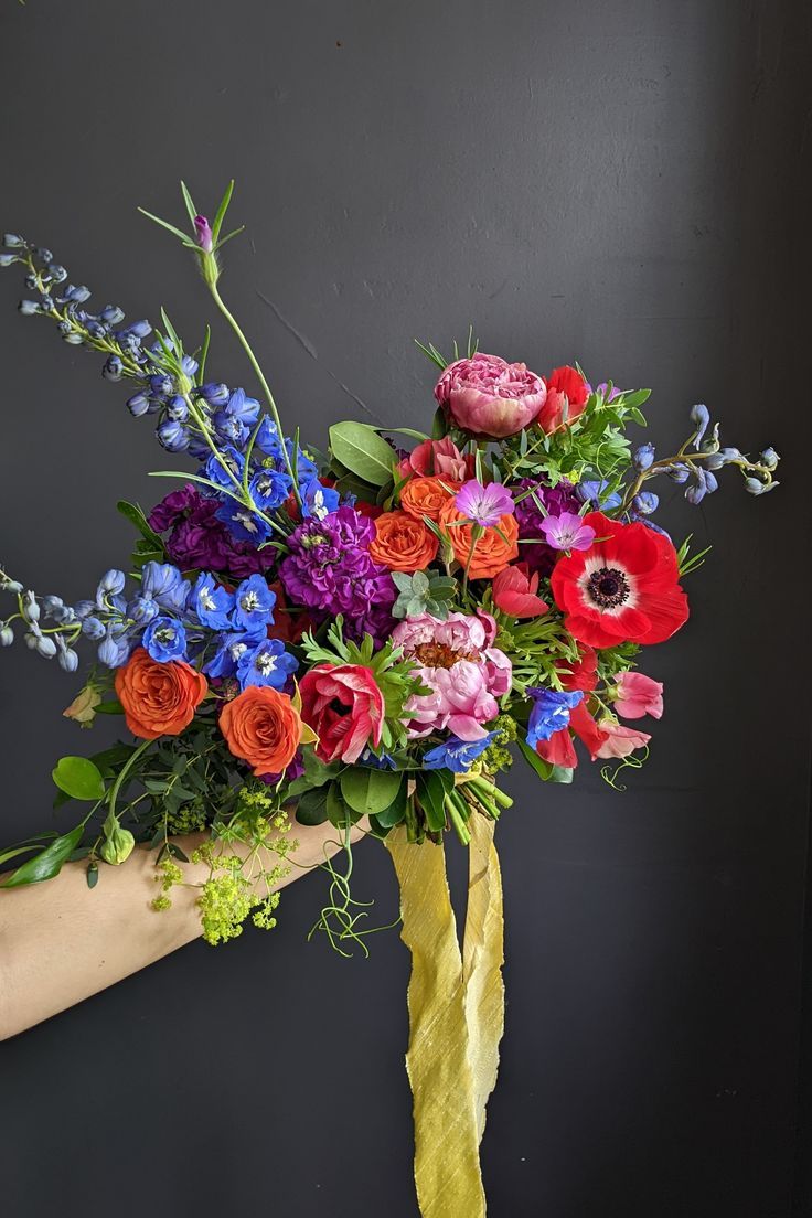 a person holding a bouquet of colorful flowers in their left hand, against a black wall