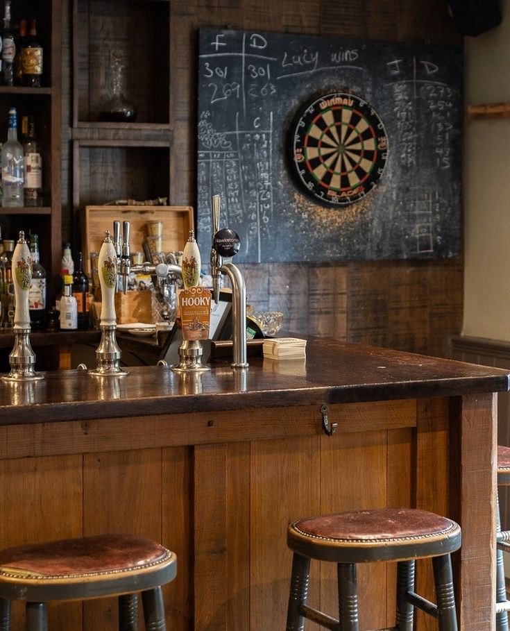 a bar with three stools in front of it and a dart board on the wall