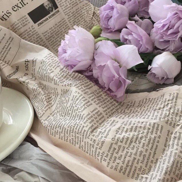 purple flowers are sitting on top of an old newspaper next to a cup of coffee