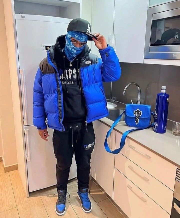 a man standing in a kitchen wearing a blue jacket and bandana covering his face