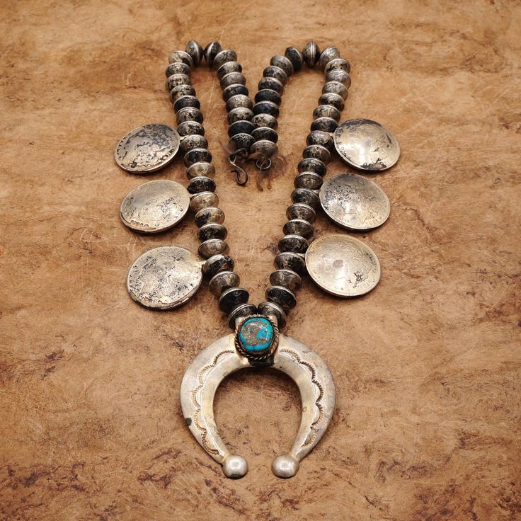 This Unique Heavy Old Pawn Squash Blossom Necklace Is A Stunning Example Of Navajo Jewelry Utilizing Mercury Dimes As Navajo Pearls And The Six Cupped Morgan Silver Dollar Coins Representing The Blossoms. The Morgan Dollars Date 1921 With The Top Left Dollar Date, 1892. The Horseshoe-Shaped Naja Pendant Features A Very Beautiful Oval-Shaped Dark Turquoise Gemstone, Set In A Bezel Style. The Pendant Hangs From A 26.5-Inch Silver Necklace, With What Appears As Hair Or Some Other Material, Along Wi Naja Necklace, Naja Pendant, Silver Dollar Coin, Turquoise Squash Blossom, Navajo Pearls, Morgan Dollars, Navajo Jewelry, Squash Blossom Necklace, Morgan Silver Dollar