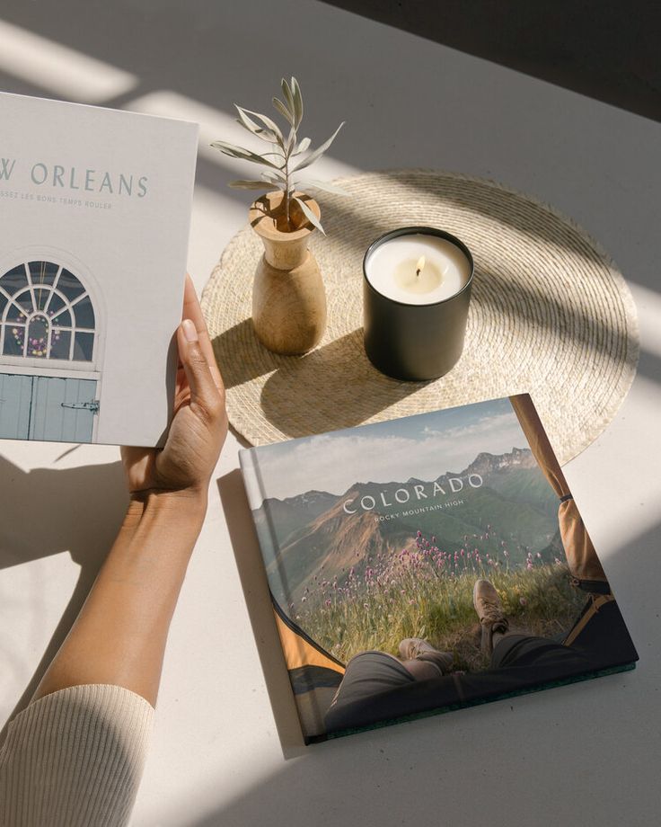 a person holding up a copy of the book colorado by tony orleans on a table next to a candle and potted plant