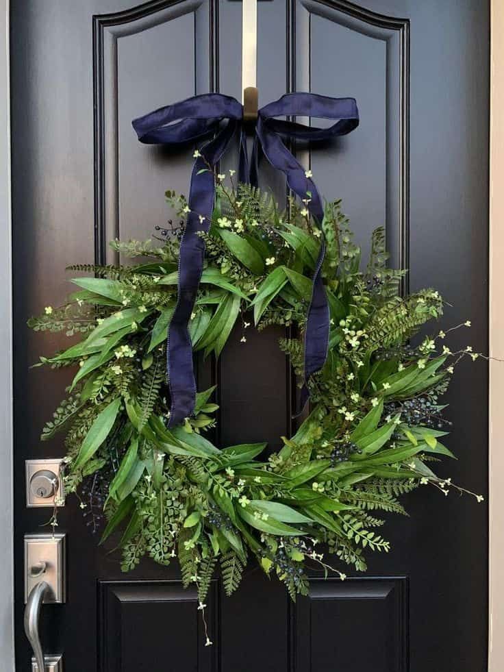 a wreath with blue ribbon hanging on the front door to a black door that is decorated with greenery