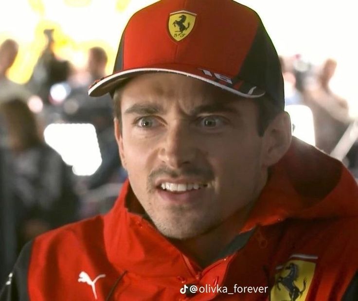 a man wearing a red and black jacket with a ferrari hat on his head is looking at the camera