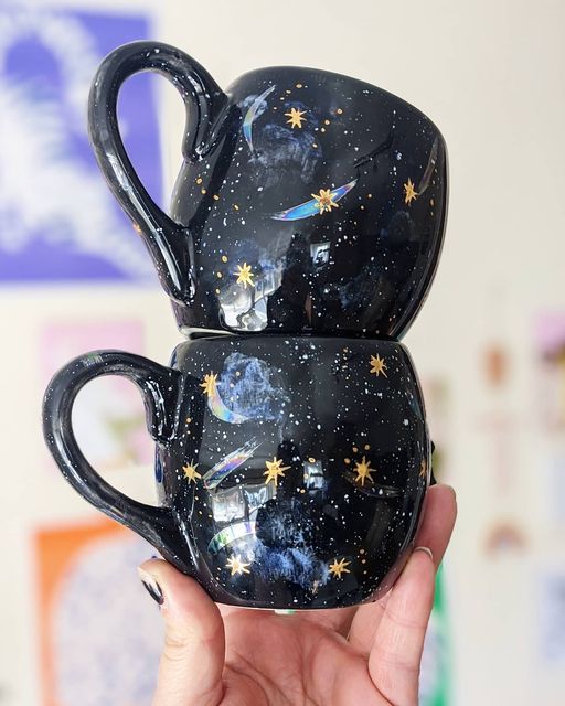 two black coffee mugs with stars painted on them are held up in the air