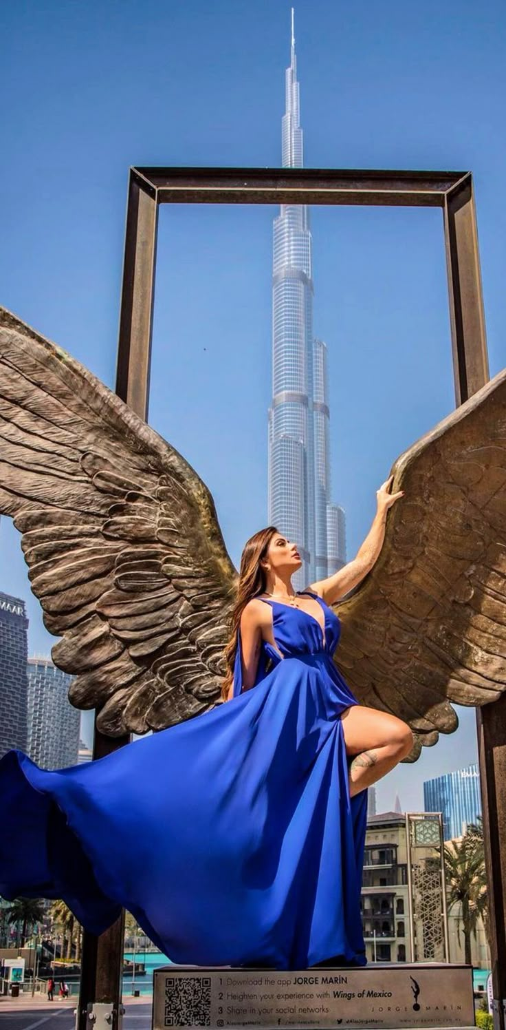 a woman in a blue dress sitting on top of an angel statue with the burj building in the background