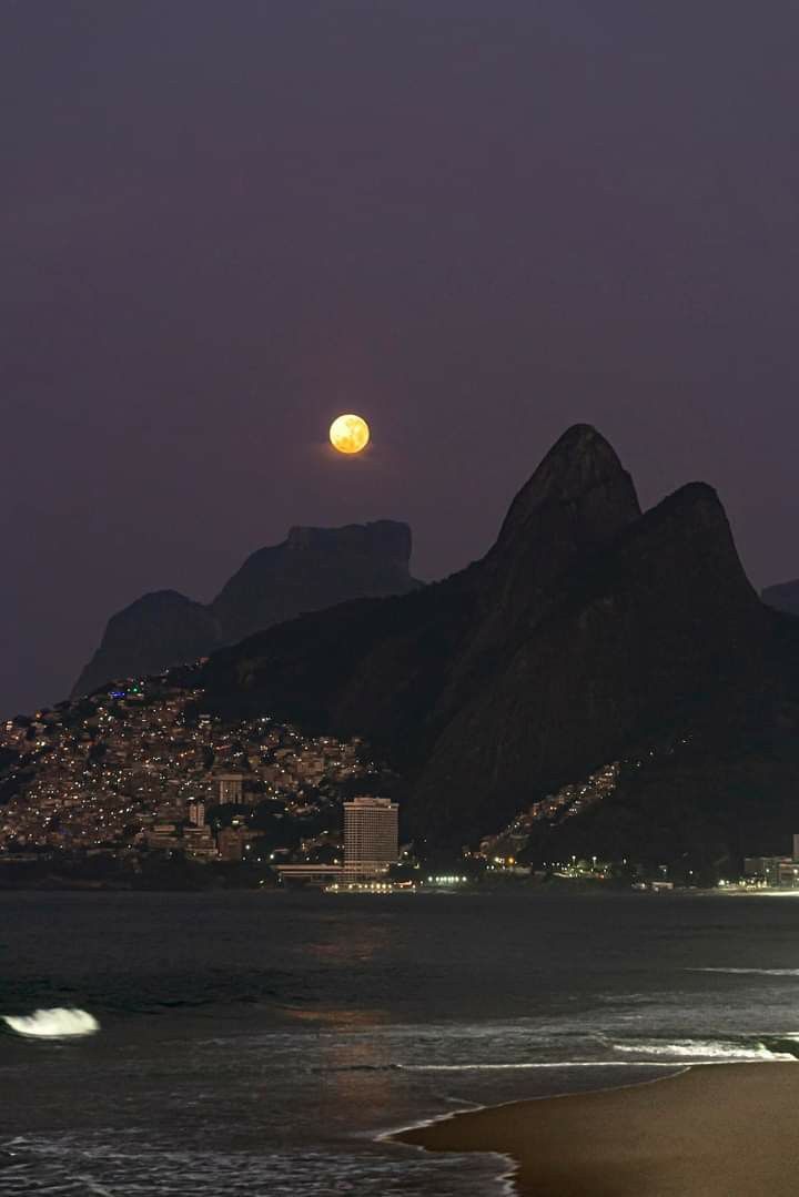 a full moon is seen over the ocean with mountains and buildings in the background at night