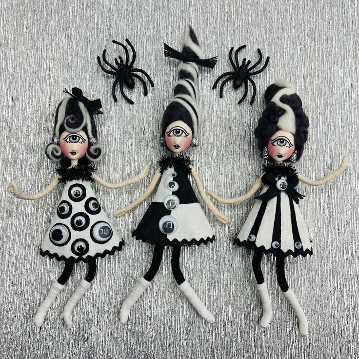 three dolls are dressed in black and white outfits with spider decorations on their heads, holding hands