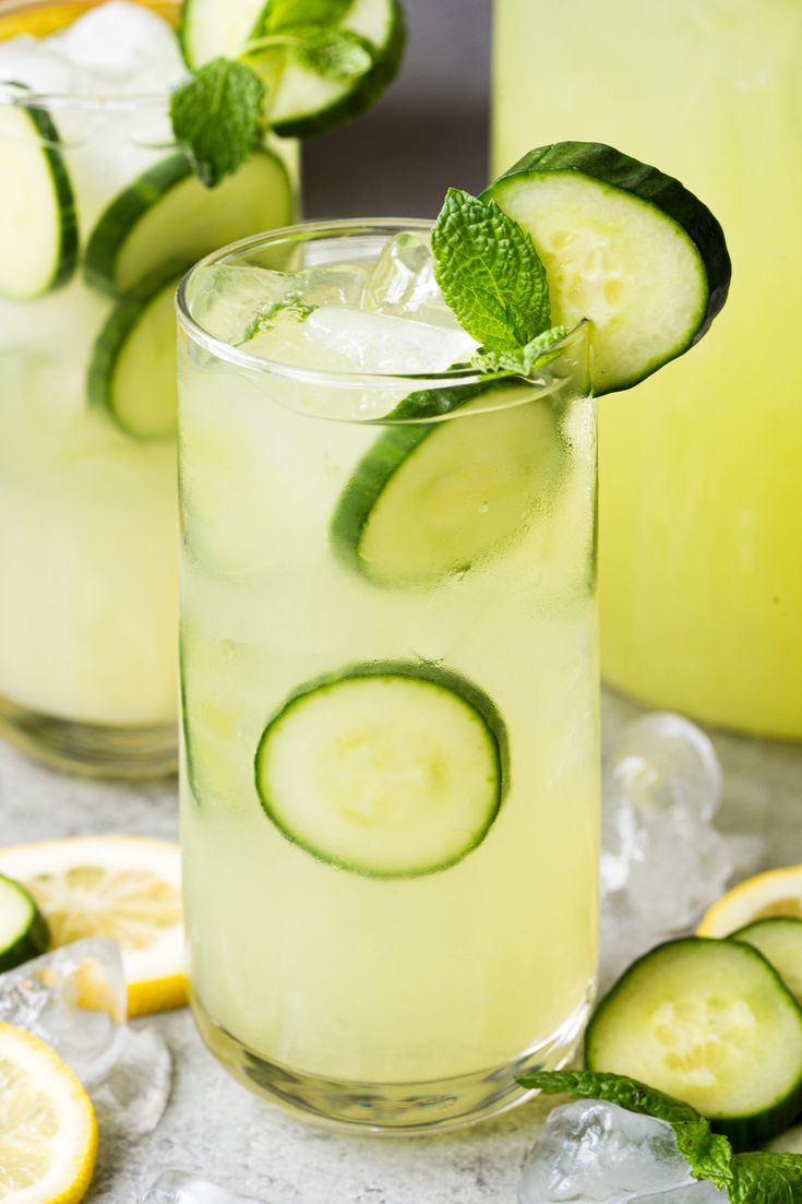 two glasses filled with lemonade and cucumber on top of a white table