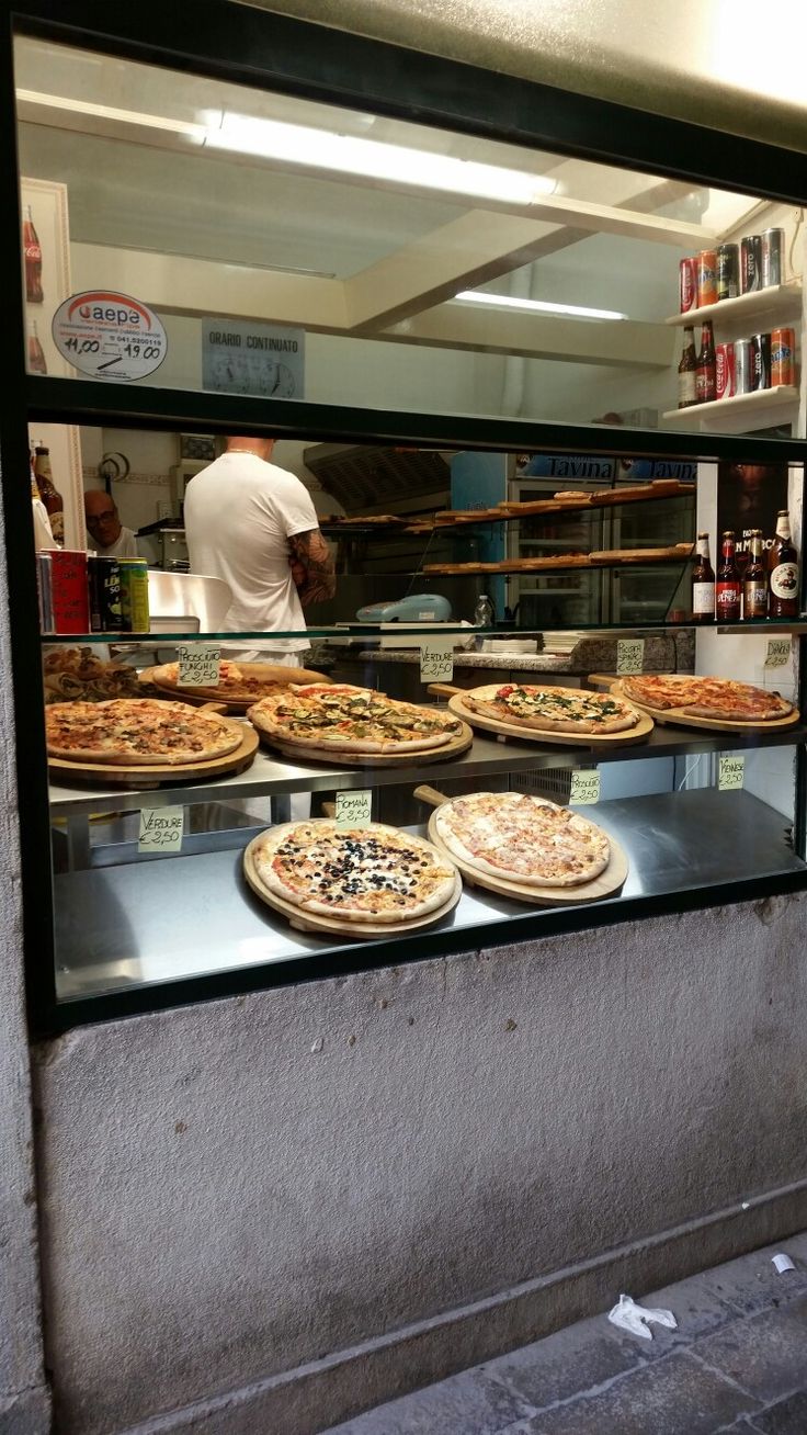 several pizzas are on display behind glass in a store window, with a man standing at the counter