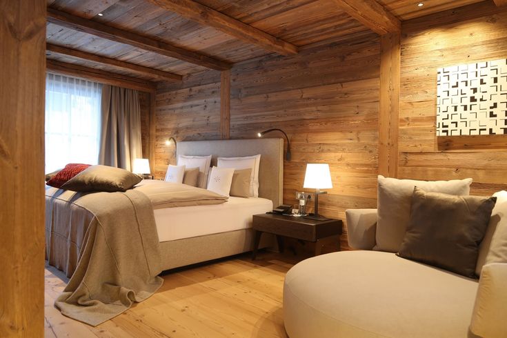 a bedroom with wooden walls and flooring has a large bed in the corner, along with two chairs