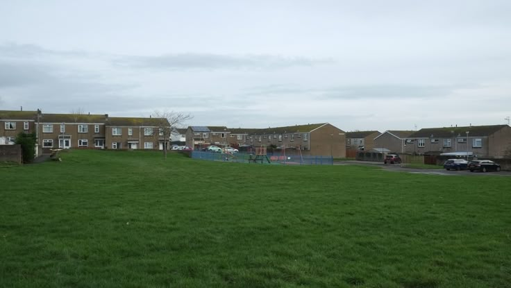 a grassy field with houses in the background and cars parked on the road behind it