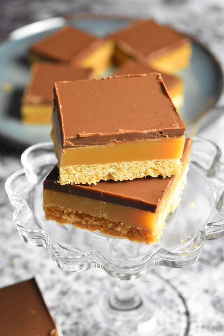 three pieces of chocolate and caramel dessert on a glass cake plate with two plates in the background