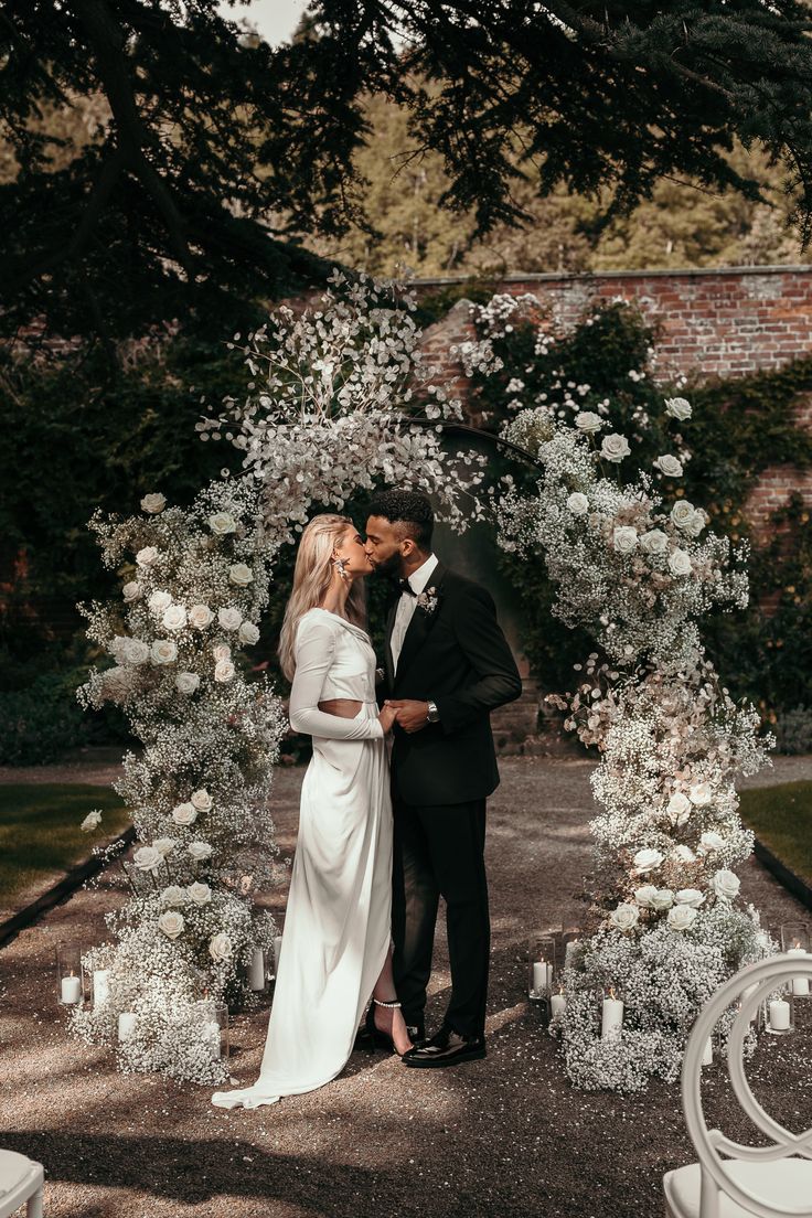 a bride and groom kissing in front of an arch with white flowers on the side