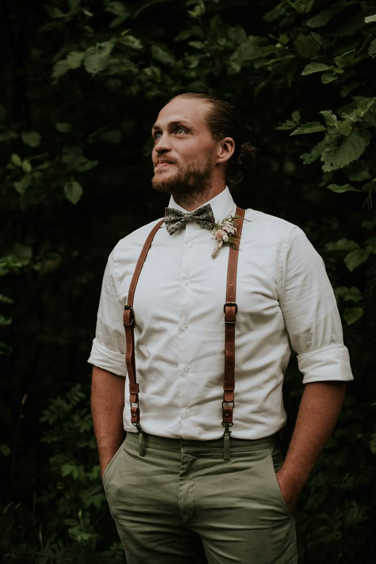 a man with a bow tie and suspenders standing in front of some green trees