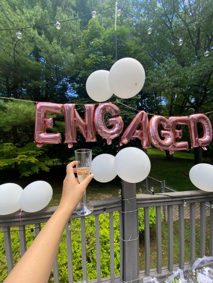 someone holding a wine glass in front of an engaged sign with balloons and streamers